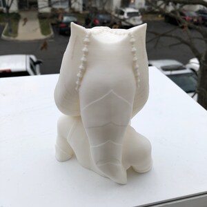 May include: White 3D printed figurine of a character with a large head and a long, pointed tail. The figurine is sitting with its legs crossed and its arms folded.