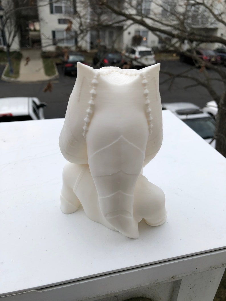 May include: White 3D printed figurine of a character with a large head and a long, pointed tail. The figurine is sitting with its legs crossed and its arms folded.