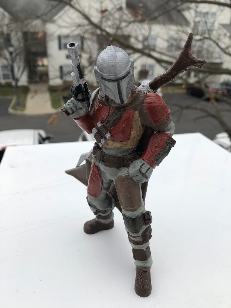 May include: A 3D printed action figure of a Mandalorian character from the Star Wars universe. The figure is wearing a gray helmet with a T-shaped visor, a red and brown armor, and holding a blaster rifle in one hand. The figure is standing on a white surface.