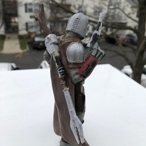 May include: A 3D printed action figure of a character wearing a brown and grey suit with a white cape and a red and grey helmet. The figure is holding a gun in one hand and a staff in the other.