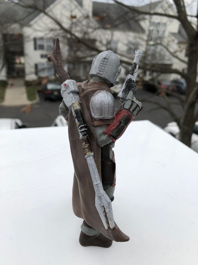 May include: A 3D printed action figure of a character wearing a brown and grey suit with a white cape and a red and grey helmet. The figure is holding a gun in one hand and a staff in the other.