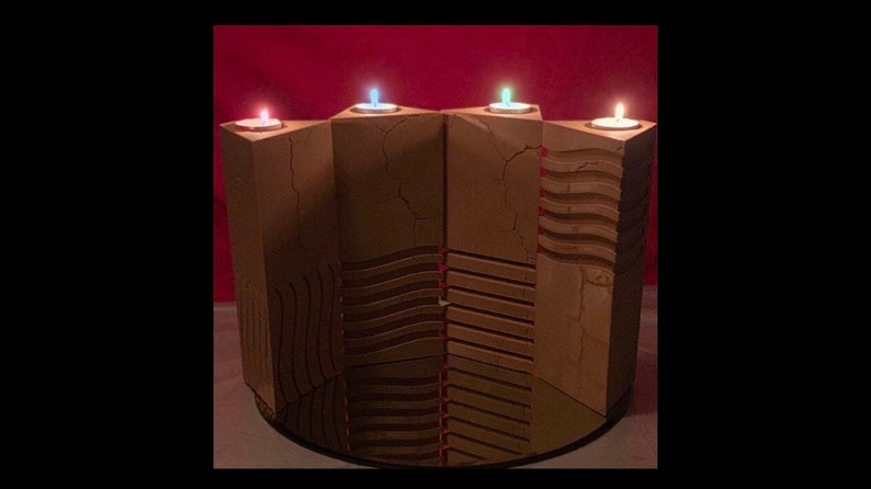 May include: A wooden candle holder with four candle slots. The holder is made of light brown wood with a textured surface. The candles are lit and are different colors: red, blue, and white.
