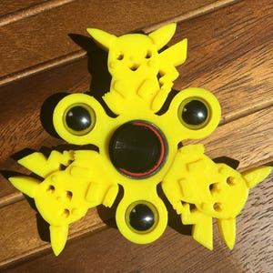 May include: A yellow fidget spinner with three Pikachu characters. The spinner has a black center with a red ring around it.
