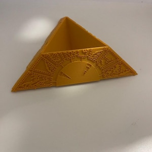 Puede incluir: Objeto decorativo triangular de color amarillo dorado con intrincados patrones en relieve. El objeto tiene un diseño similar a un reloj en su cara frontal y es probablemente un artículo decorativo o un contenedor.