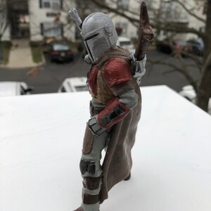 May include: A 3D printed action figure of a Mandalorian character from the Star Wars franchise. The figure is wearing a grey helmet, brown armor, and a brown cape. The figure is holding a weapon in its right hand.