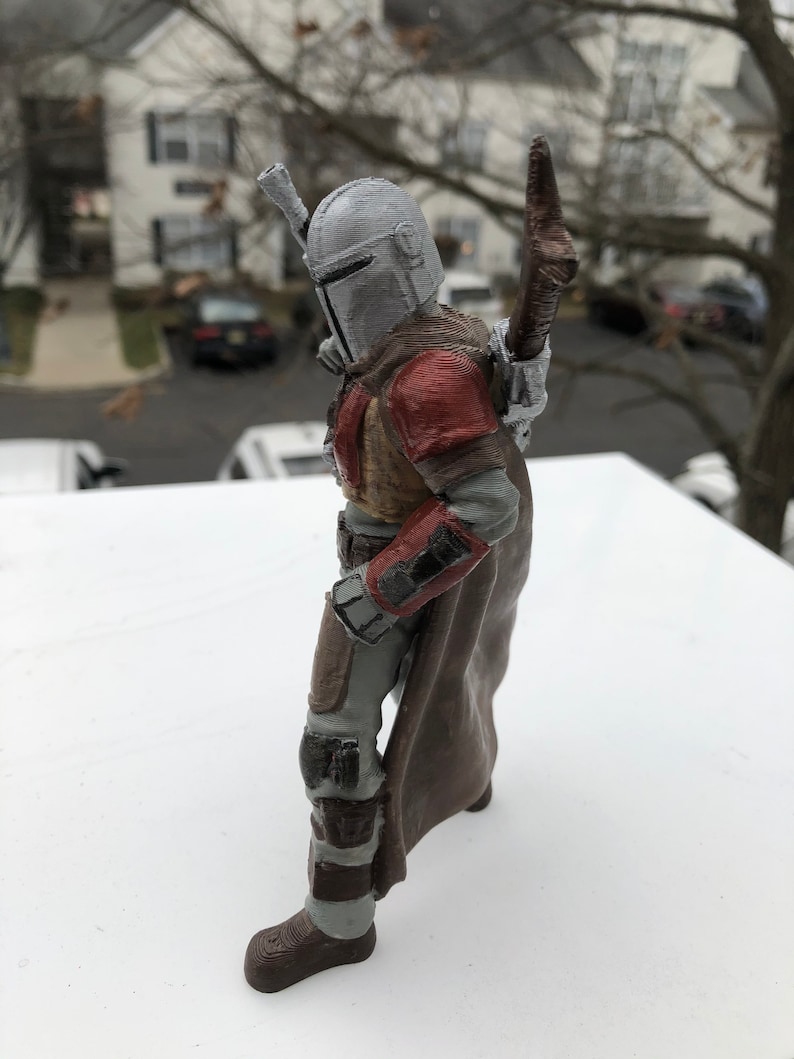 May include: A 3D printed action figure of a Mandalorian character from the Star Wars franchise. The figure is wearing a grey helmet, brown armor, and a brown cape. The figure is holding a weapon in its right hand.