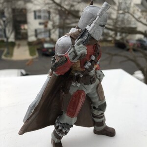 May include: A 3D printed action figure of a Mandalorian warrior. The figure is wearing a brown and gray armor with red accents. The figure is holding a blaster rifle in its right hand. The figure is standing on a white surface.