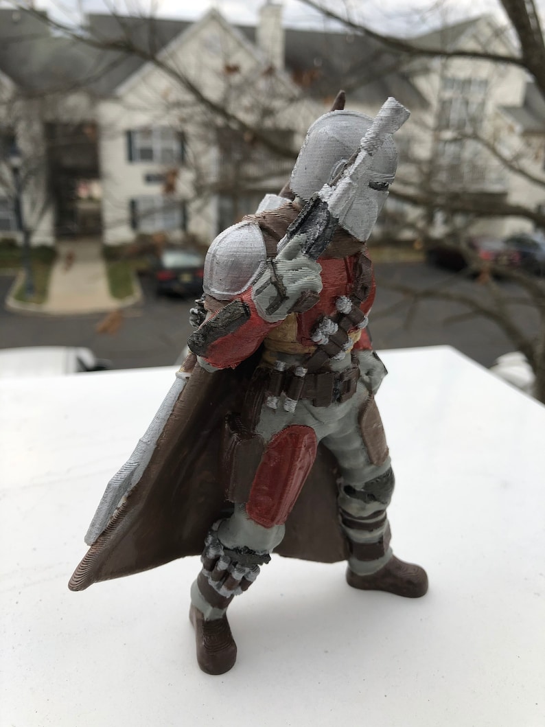 May include: A 3D printed action figure of a Mandalorian warrior. The figure is wearing a brown and gray armor with red accents. The figure is holding a blaster rifle in its right hand. The figure is standing on a white surface.
