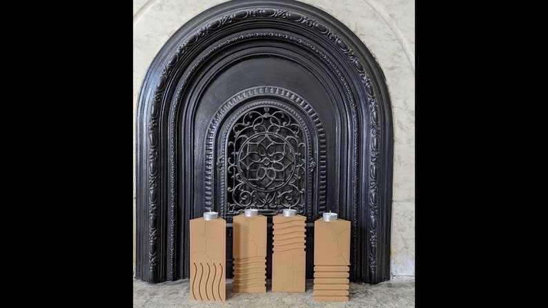 May include: Four brown wooden candle holders with different carved designs, arranged in a row in front of a black fireplace.