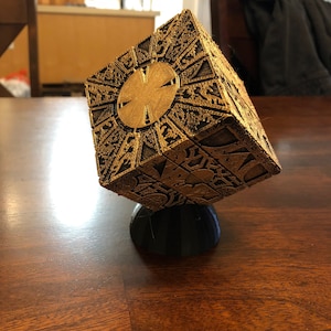 May include: A gold-colored, intricately detailed cube puzzle with a circular design on one side. The cube is sitting on a black base.