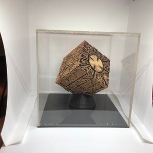 May include: A detailed, gold-colored puzzle box with intricate patterns and a central circular design, displayed inside a clear acrylic case. The cube rests on a black, rounded base, set against a white background.