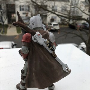 May include: A detailed action figure of a character wearing a brown cape and helmet with a silver and red chest plate. The figure is holding a weapon in its right hand.