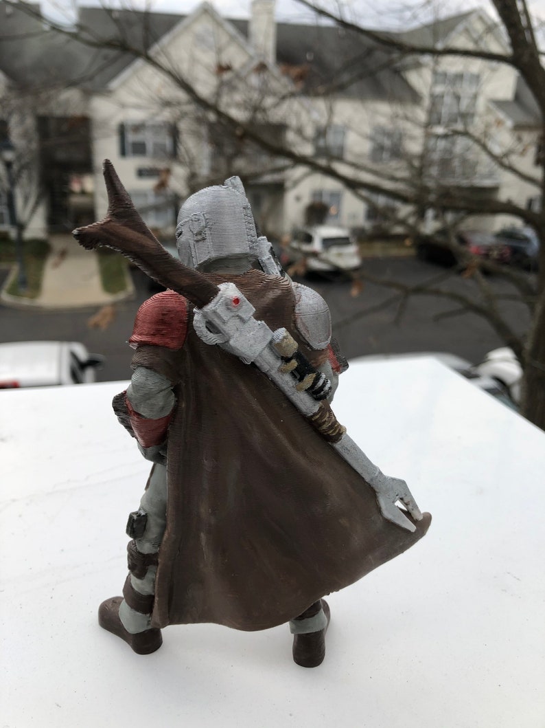 May include: A detailed action figure of a character wearing a brown cape and helmet with a silver and red chest plate. The figure is holding a weapon in its right hand.