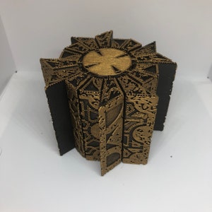 May include: A gold-colored metal puzzle box with intricate designs. The box is closed and has a star-shaped top.