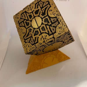 May include: A gold-colored, intricate, cube-shaped puzzle box with black detailing sits on a smaller, triangular, gold-colored base. The box has a complex, geometric pattern on all sides.