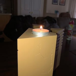 May include: A lit candle in a small, round, silver holder sits on top of a tall, rectangular, beige candle holder with a cracked surface.