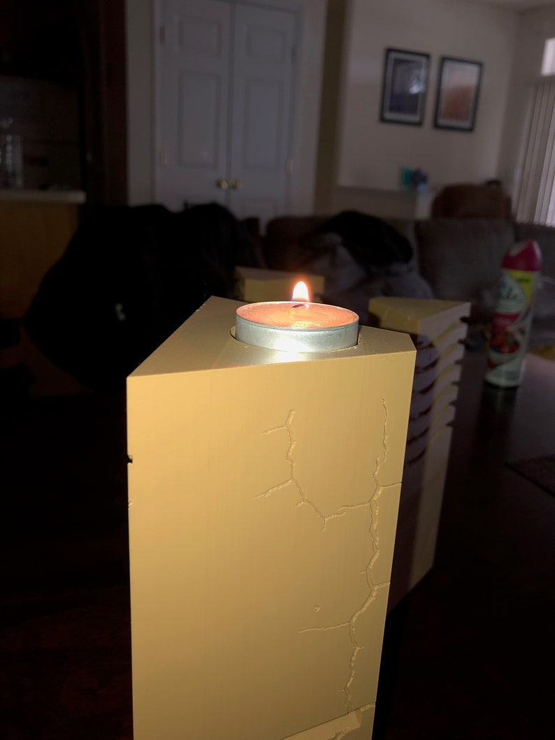 May include: A lit candle in a small, round, silver holder sits on top of a tall, rectangular, beige candle holder with a cracked surface.
