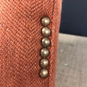 May include: Detail of a rust-coloured blazer sleeve. The fabric has a textured, woven appearance. Six bronze-coloured, round buttons are vertically aligned. The blazer's colour is a warm, earthy tone, similar to terracotta.