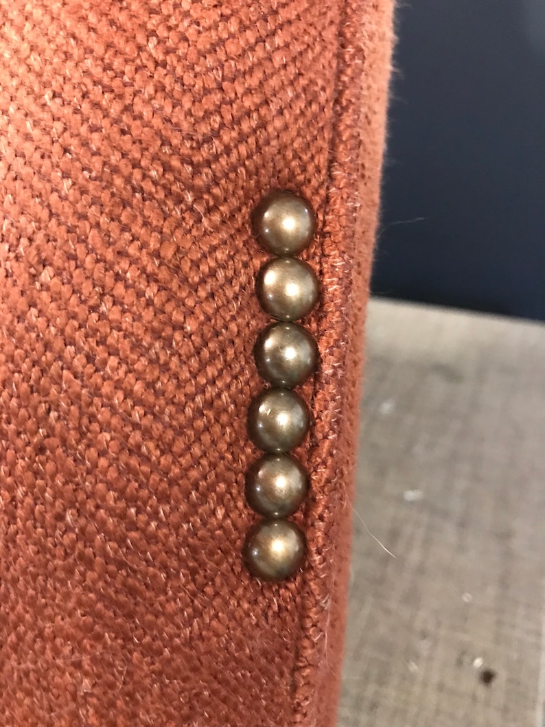 May include: Detail of a rust-coloured blazer sleeve. The fabric has a textured, woven appearance. Six bronze-coloured, round buttons are vertically aligned. The blazer's colour is a warm, earthy tone, similar to terracotta.