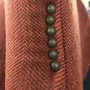 May include: Close-up of a rust-coloured jacket with a herringbone weave. Six dark, round buttons are visible along the edge of the jacket. The fabric has a textured appearance.