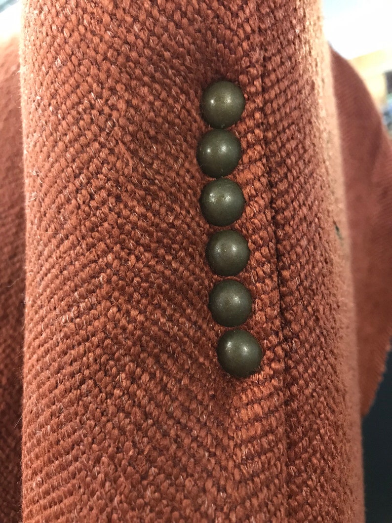 May include: Close-up of a rust-coloured jacket with a herringbone weave. Six dark, round buttons are visible along the edge of the jacket. The fabric has a textured appearance.