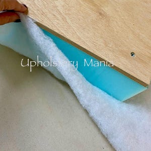 May include: A white fabric is being pulled back to reveal a piece of blue foam and a piece of wood with the text "Upholstery Mania" printed on it.