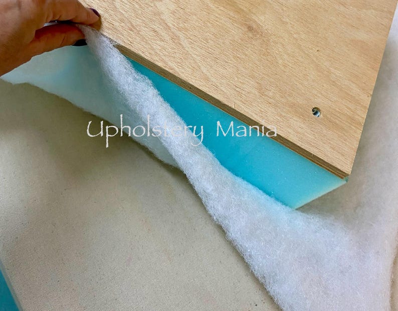 May include: A white fabric is being pulled back to reveal a piece of blue foam and a piece of wood with the text "Upholstery Mania" printed on it.