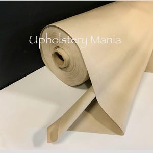 May include: A roll of beige upholstery fabric. The fabric is rolled up and the end of the roll is unfurled. The fabric is smooth and has a slight sheen. The text "Upholstery Mania" is visible on the fabric.
