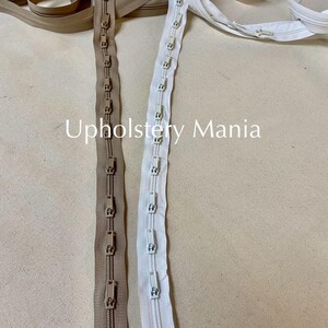 May include: Two zipper tapes, one beige and one white, with metal sliders and plastic teeth. The zipper tapes are laid out on a beige fabric surface. The text "Upholstery Mania" is visible in the image.