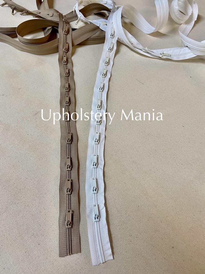 May include: Two zipper tapes, one beige and one white, with metal sliders and plastic teeth. The zipper tapes are laid out on a beige fabric surface. The text "Upholstery Mania" is visible in the image.