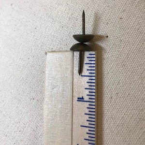 May include: Two antique brass upholstery tacks with rounded heads and sharp shanks, next to a ruler. The tacks measure approximately 2.5 cm in height. The image shows a close-up of the tacks and ruler.