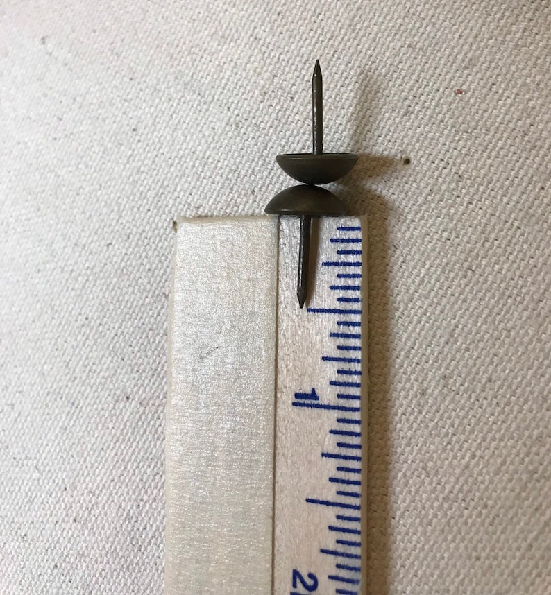May include: Two antique brass upholstery tacks with rounded heads and sharp shanks, next to a ruler. The tacks measure approximately 2.5 cm in height. The image shows a close-up of the tacks and ruler.