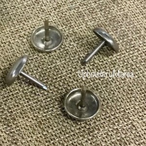 May include: Four silver upholstery tacks with a round head and a pointed nail. The tacks are on a brown fabric surface.
