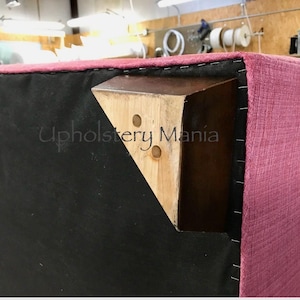 May include: Close-up of a pink upholstered sofa with a wooden leg. The leg is attached to the sofa with black staples. The text "Upholstery Mania" is visible on the leg.