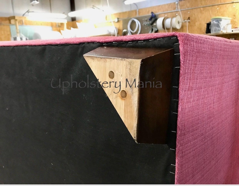 May include: Close-up of a pink upholstered sofa with a wooden leg. The leg is attached to the sofa with black staples. The text "Upholstery Mania" is visible on the leg.