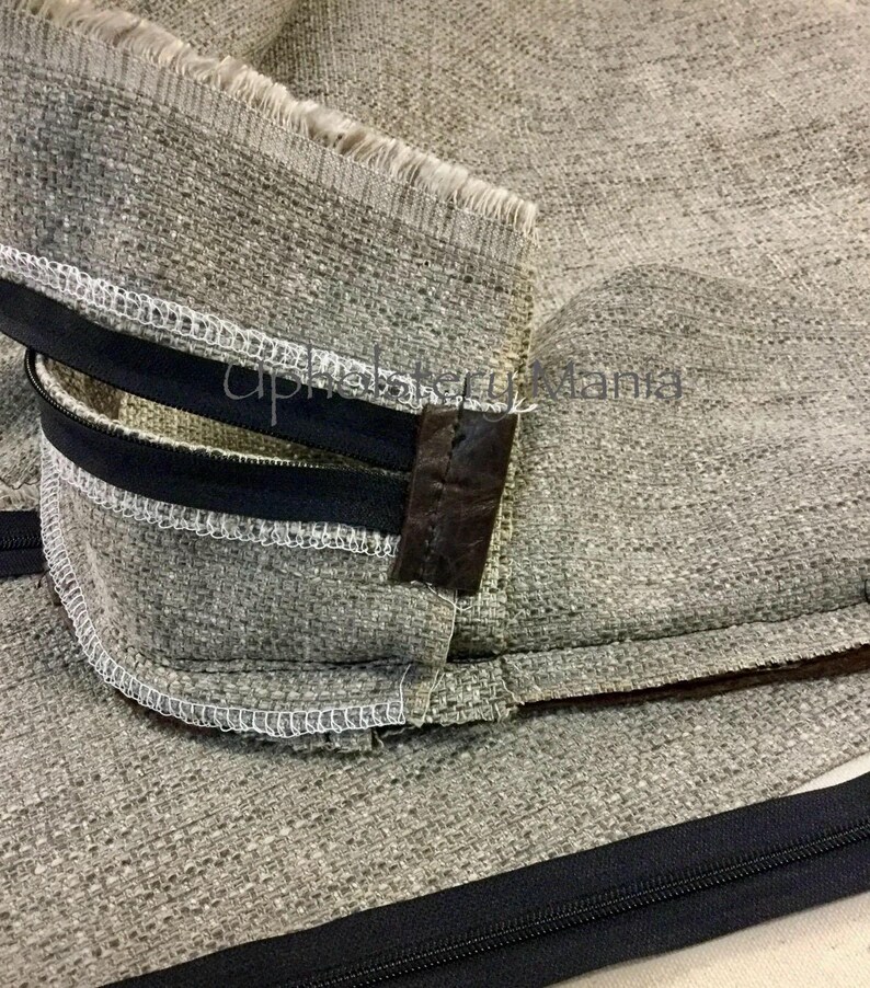 May include: Close-up of a gray fabric with a black zipper and brown leather trim. The fabric is sewn with white thread.