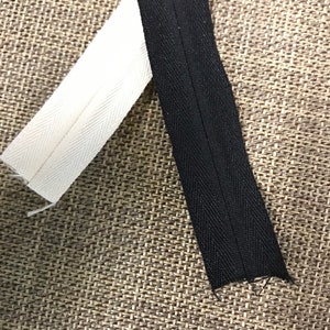 May include: Two pieces of zipper tape, one white and one black, are laid out on a brown textured surface. The white tape has a herringbone pattern and the black tape has a straight stitch.
