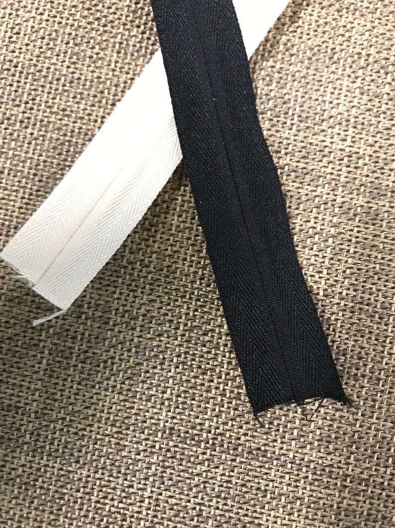 May include: Two pieces of zipper tape, one white and one black, are laid out on a brown textured surface. The white tape has a herringbone pattern and the black tape has a straight stitch.