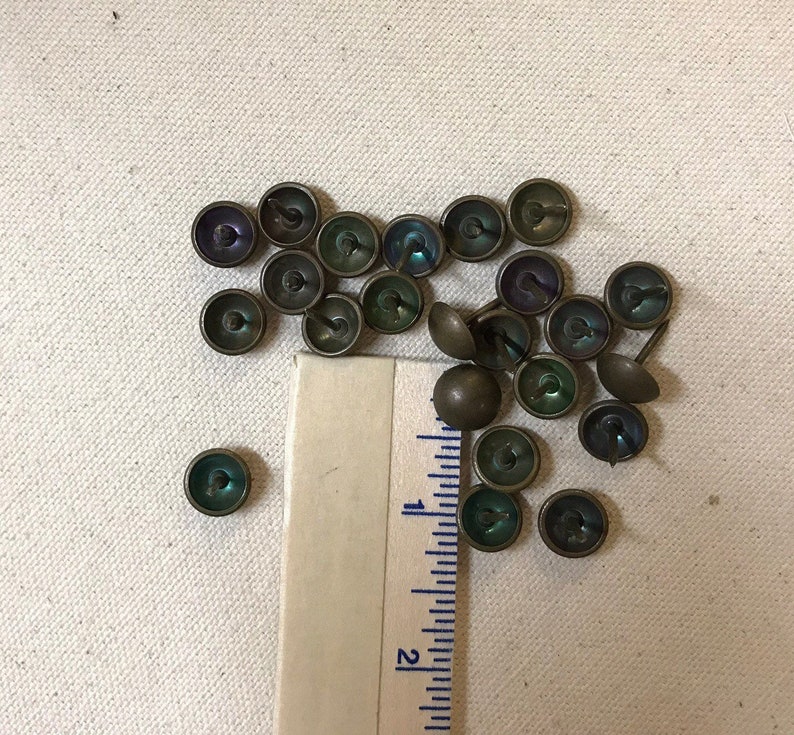 May include: A selection of antique upholstery tacks with iridescent green and purple centers and bronze-coloured metal caps. A ruler is placed next to the tacks. The tacks are small, round, and have a domed head.