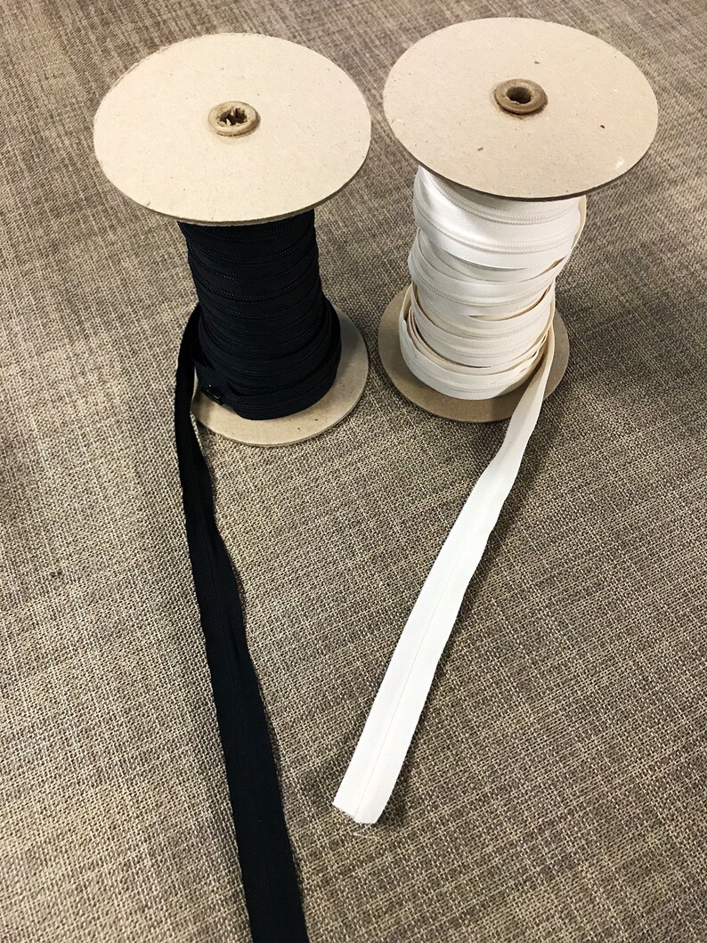May include: Two spools of zipper tape, one black and one white, are wound around cardboard spools. The spools are sitting on a brown fabric surface.