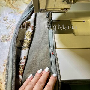 May include: A close-up of a sewing machine needle stitching a zipper into a fabric cushion. The fabric is a light blue with a floral pattern and the zipper is a light blue. A hand is holding the fabric down.
