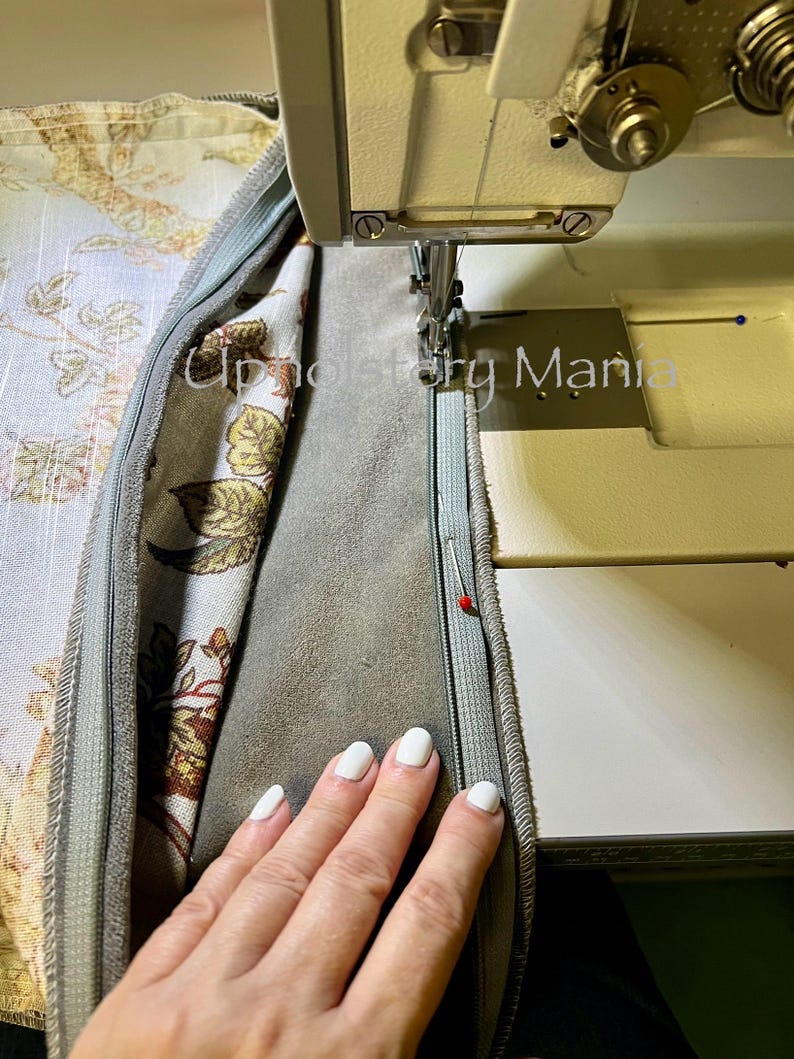 May include: A close-up of a sewing machine needle stitching a zipper into a fabric cushion. The fabric is a light blue with a floral pattern and the zipper is a light blue. A hand is holding the fabric down.