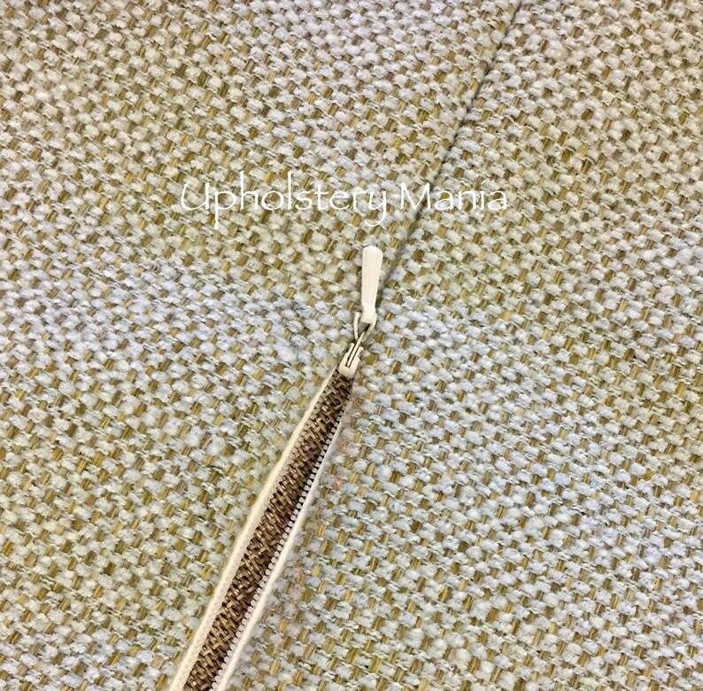 May include: Close-up of a zipper on a beige fabric cushion cover. The zipper is metal and has a white pull tab.
