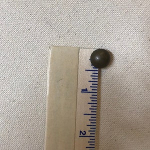 May include: A small, round, bronze-coloured upholstery tack sits next to a wooden ruler. The ruler is marked with inch measurements. The tack is approximately 1.3 cm in diameter. The background is a light beige fabric.