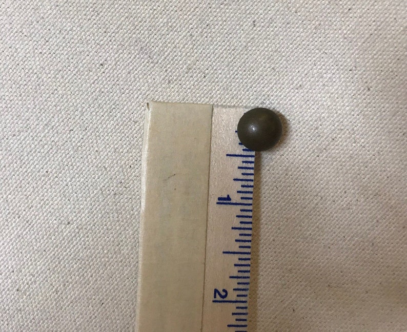 May include: A small, round, bronze-coloured upholstery tack sits next to a wooden ruler. The ruler is marked with inch measurements. The tack is approximately 1.3 cm in diameter. The background is a light beige fabric.