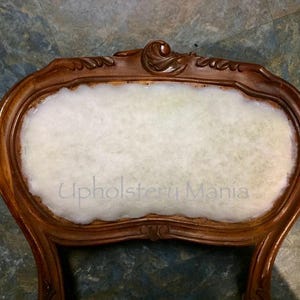 May include: Close-up of the back of a wooden chair with a white cotton padding. The chair has a carved design on the top and is in the process of being reupholstered. The text "Upholstery Mania" is visible on the padding.