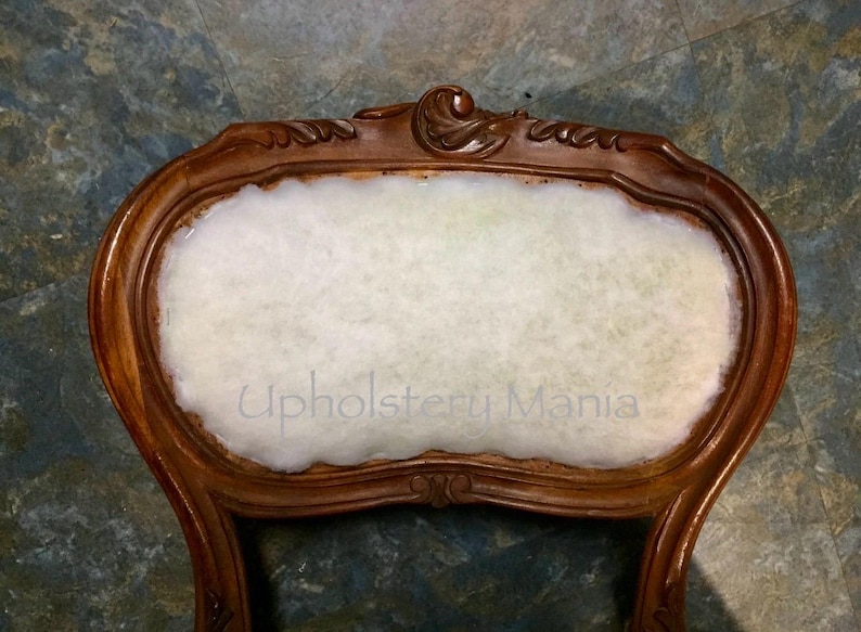 May include: Close-up of the back of a wooden chair with a white cotton padding. The chair has a carved design on the top and is in the process of being reupholstered. The text "Upholstery Mania" is visible on the padding.