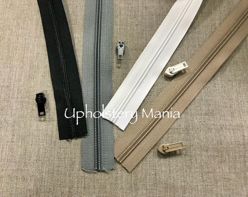 May include: Four different types of zipper tape, including black, gray, white, and beige. The zipper tape is laid out on a beige fabric background. The text "Upholstery Mama" is visible in the center of the image.