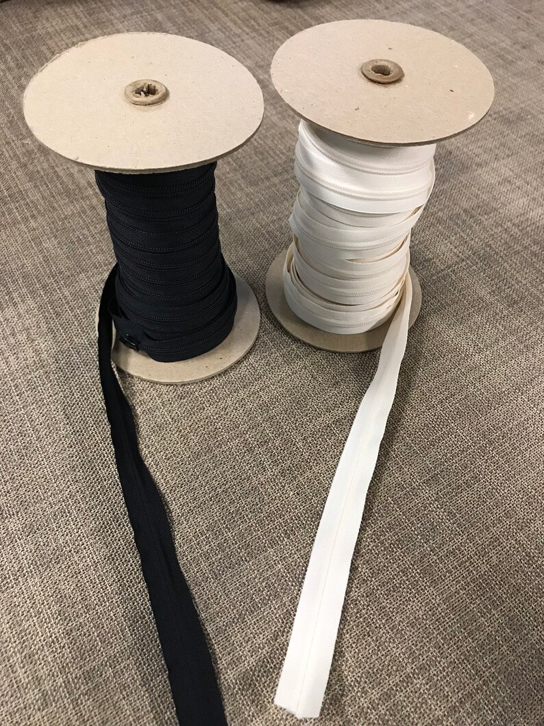 May include: Two spools of zipper tape, one black and one white, are unwinding on cardboard spools. The zipper tape is laying on a brown textured surface.