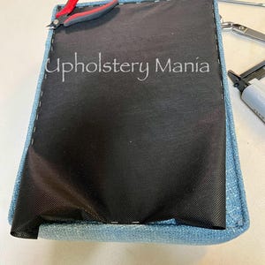 May include: A close-up of a blue fabric-covered cushion with a black fabric lining. The cushion is being stapled together with a staple gun. The text "Upholstery Mania" is visible on the cushion.